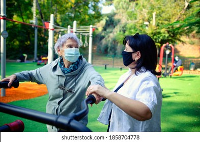 A Professional Caregiver Helps A Senior Adult Woman To Use A Fitness Outdoor Machine Properly. Both Wearing Face Medical Masks Due To The Coronavirus Pandemic.