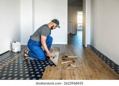 Professional carpenter master worker in blue overalls joining vinyl or laminate floor covering at home renovation. Industrial theme