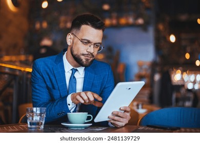 Professional Caucasian male in a sharp blue suit uses a digital tablet, managing tasks while enjoying a coffee break indoors. - Powered by Shutterstock