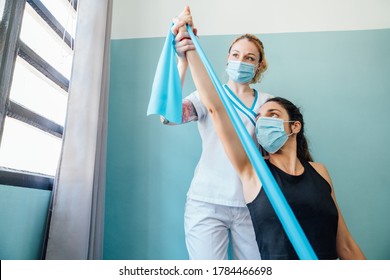Professional Caucasian physical therapy woman at a clinic giving a Pilates stretch treatment with a rubber band to a client with a face mask due to the covid 19 coronavirus pandemic. - Shutterstock ID 1784466698