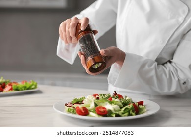 Professional chef adding pepper to delicious salad at white marble table in kitchen, closeup