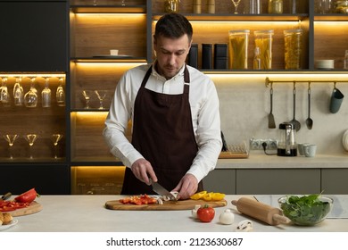 Professional chef in apron tells to camera about cooking delicious dish with vegetables. Young man blogger cuts fresh bell pepper on board at table in kitchen - Powered by Shutterstock