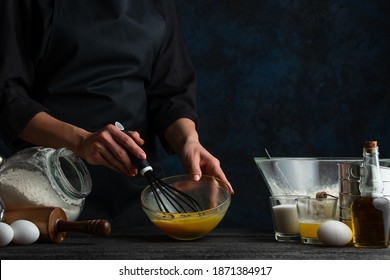 Professional Chef Beats Eggs In The Glass Bowl For Preparing The Dough. Ingredients For Cooking On The Background. Dark Blue Background Space. Frozen Motion. Cooking Process.