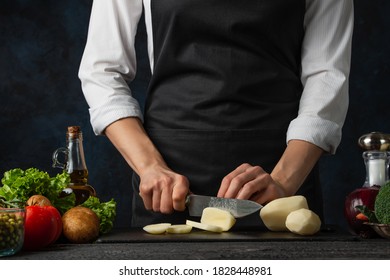 The professional chef in black apron cuts with knife potato on black chopped board on dark blue background. Backstage of preparing restaurant meal for dinner. Food concept. Close-up.