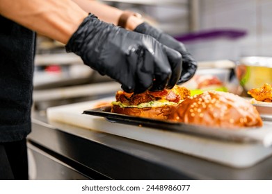 Professional chef in black gloves assembling a juicy burger in a kitchen. Focus on hands and burger with visible layers of lettuce, cheese, and sauce - Powered by Shutterstock