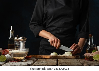 The professional chef in black uniform cuts with knife whole white onion on chopped wooden board at rustic table with ingredients for cooking background. Backstage of preparing meal with vegetables. - Powered by Shutterstock