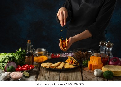 Professional chef in black uniform pours spices on corn tortilla for cooking tacos on dark blue background. Traditional mexican cuisine. Concept of tasty street food. Frozen motion.