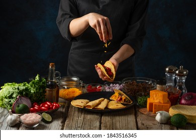 Professional chef in black uniform pours cheese on corn tortilla for cooking tacos on dark blue background. Traditional mexican cuisine. Concept of tasty street food. Frozen motion.