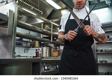 Professional Chef. Brutal Young Man In Apron With Several Tattoos On His Arms Sharpening A Knife In A Restaurant Kitchen