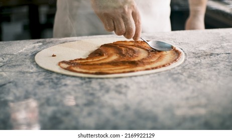 Professional Chef Is Cooking Pizza, Applying Delicious Tomato Sauce On Dough With A Spoon. Pizza Cooker Spreading The Sauce. 