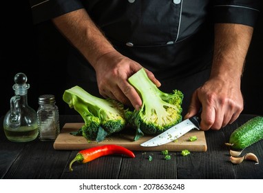 Professional chef cuts fresh Broccoli with a knife for salad on vintage kitchen table with fresh vegetables. Cooking and restaurant or cafe concept