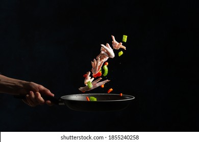 The Professional Chef Hand Toss Up Raw Chicken Legs With Chopped Vegetables Above The Frying Pan On Black Background. Backstage Of Cooking Grilled Meat For Dinner. Frozen Motion. Asian Cuisine.