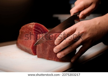 Professional chef neatly cutting part of fresh raw tuna with sharp Japanese knife in restaraunt kitchen. Dark blurred background. Asian food restaraunt