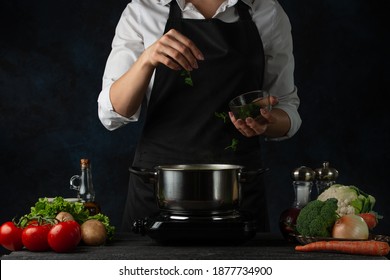 Professional chef pours parsley into pan for cooking soup on dark blue background. Backstage of preparing meal. Healthy dish. Food concept. Frozen motion. Cookbook.
