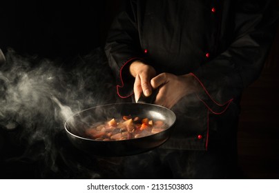 Professional chef prepares food in a frying pan with steam on a black background. The concept of restaurant and hotel service. Asian cuisine.