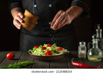 Professional chef sprinkles salt to salad of fresh vegetables on wooden table. Preparing healthy food on the kitchen.