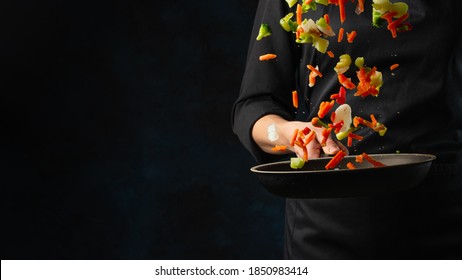 Professional Chef Throws Up Frying Mix Of Colored Vegetables Above The Pan On Dark Blue Background. Backstage Of Cooking Meal. Frozen Motion. Food Banner Concept.