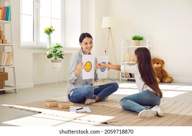 Professional children's psychologist talking to little kid about emotions. Girl chooses happy smiley emoticon from two EQ cards that female therapist shows her during interview appointment meeting