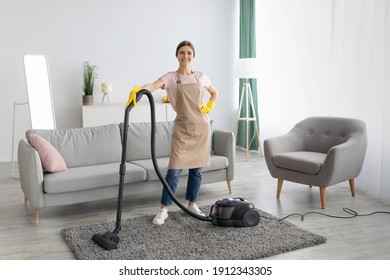 Professional Cleaning Service. Positive Young Lady In Apron And Rubber Gloves Vacuuming Living Room Of Modern House. Full Length Portrait Of Millennial Maid Tidying Apartment With Hoover