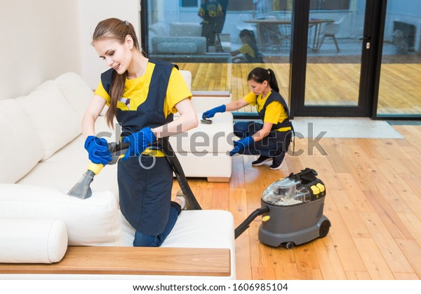 professional cleaning service. Two women in working\
uniform, in aprons. Two women in working uniform, in aprons, divide\
the cleaning of private house, cottage. Washing , vacuum cleaning\
the sofa