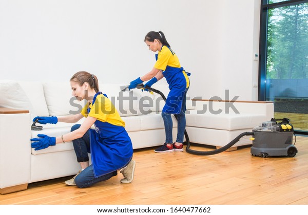 professional cleaning service. Two women in working\
uniform, in aprons. Two women in working uniform, in aprons, divide\
the cleaning of private house, cottage. Washing , vacuum cleaning\
the sofa