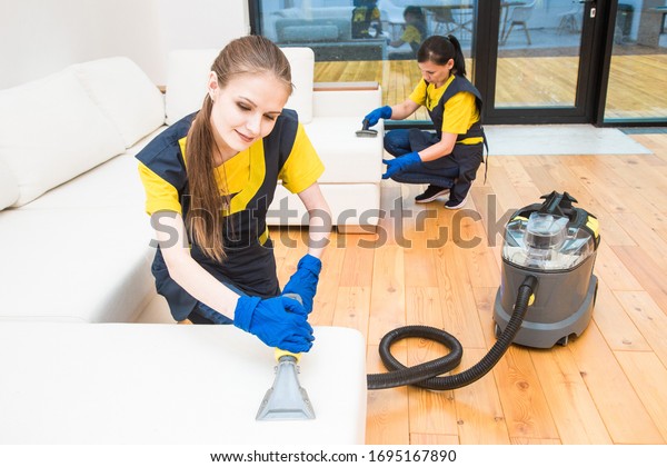 professional cleaning service. Two women in working
uniform, in aprons. Two women in working uniform, in aprons, divide
the cleaning of private house, cottage. Washing , vacuum cleaning
the sofa