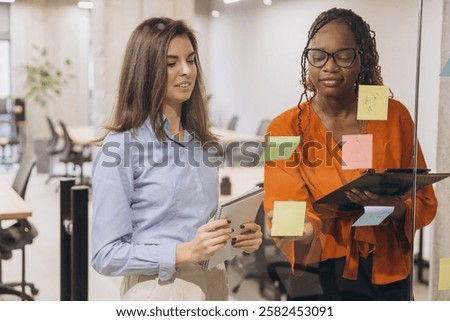 Professional colleagues collaborating, writing notes on glass wall, sharing innovative ideas in modern workspace, demonstrating strategic teamwork