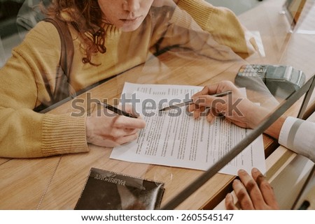 Professional completing documents at an office desk with glass partition, capturing hands and paperwork, person in yellow sweater holding pen while filling out form