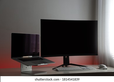 Professional computer workstation ideal for the home office consisting of a gray laptop on a stand, a large curved and black monitor, a white keyboard and a white optical mouse on a white table.