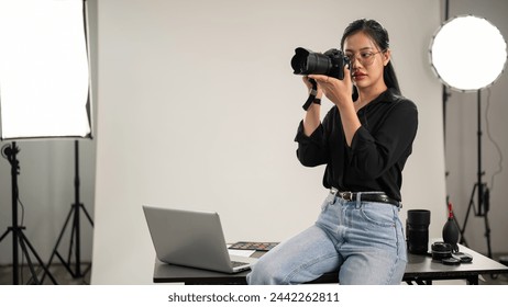 A professional, confident Asian female photographer is taking photos, holding a camera near her face, adjusting her camera, working in a studio with professional lighting equipment. - Powered by Shutterstock