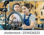 Professional confident young female bicycle technician adjusting bike gear system, fixing sprockets on rear wheel using cassette pliers and explaining repair details to female client in workshop