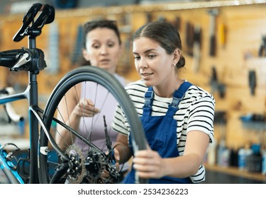 Professional confident young female bicycle technician adjusting bike gear system, fixing sprockets on rear wheel using cassette pliers and explaining repair details to female client in workshop - Powered by Shutterstock