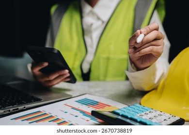 Professional Construction Worker Analyzing Data on Smartphone with Charts and Calculator on Table.