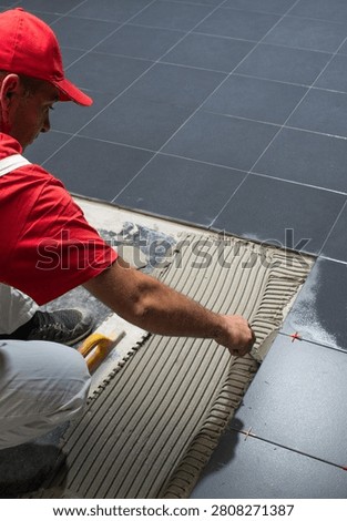 Professional construction worker installing large dark ceramic floor tiles using tile adhesive and a notched trowel. Interior flooring renovation with precision tile spacers and leveling tools, showcasing craftsmanship, home improvement, and modern construction.
