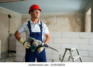 Professional construction worker in uniform standing with rotary hammer drill. Portrait of contractor in hardhat and overalls posing with jackhammer near step ladder and masonry indoors.