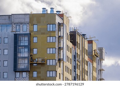 Professional construction workers are actively cladding a modern multi-story residential building, applying insulation material and installing exterior facade elements using a suspended platform