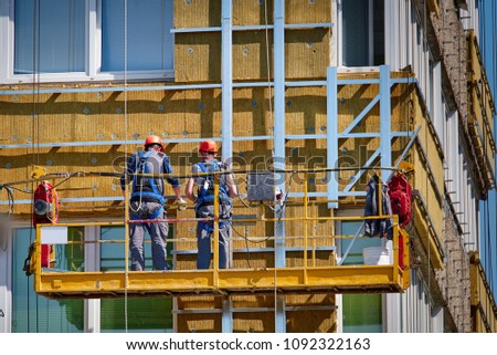 Professional construction workers on the scaffold elevator insulated wall facade with mineral wool. roofers wearing safety harness insulating wall facade at height on old building in a crane. 