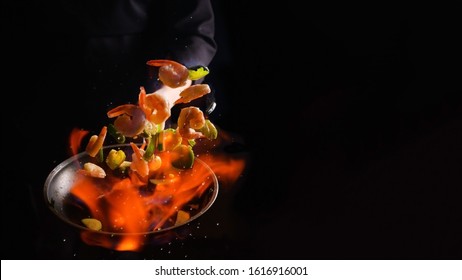 Professional Cook Cooks Shrimps With Vegetables On Fire. Cooking Seafood, Healthy Vegetarian Food, Roasting Over An Open Fire. On A Dark Background. Hotel Service, Oriental Cuisine Asia Food