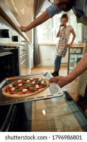 Professional Cook Making Pizza At Home. Man Putting Raw Pizza In Modern Oven For Baking. Woman Standing In The Background. Hobby, Lifestyle. Selective Focus. Vertical Shot