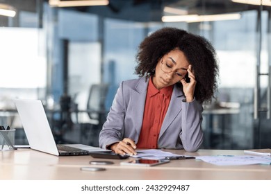 A professional corporate setting capturing a businesswoman at her desk surrounded by documents, a laptop, and a smartphone, seemingly dealing with a stressful workload. - Powered by Shutterstock