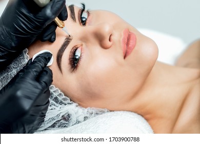 Professional cosmetologist hands are doing permanent makeup on eyebrows of young woman, close up.