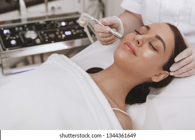 Professional cosmetologit performing microdermabrasion procedure on the face of a female client. Beautiful young woman getting face skin exfoliation treatment, copy space