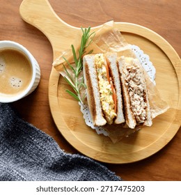 A professional culinary set featuring two types of toasted club sandwiches: a creamy egg salad sandwich with fresh tomato slices and a savory tuna salad sandwich. The golden-brown toasted bread is perfectly layered with rich fillings and garnished with a fresh rosemary sprig. Presented on a rustic wooden pizza-style serving board with white lace paper doilies and brown parchment paper. The collection includes a top-down flat lay and a 45-degree angle close-up shot, accompanied by a cup of hot espresso coffee with rich crema. Set against a wooden tabletop with a cozy grey knitted background. Ideal for cafe menus, breakfast promotion, healthy lunch blogs, and gourmet recipe content. Bright, natural lighting highlights the fresh textures and appetizing presentation.