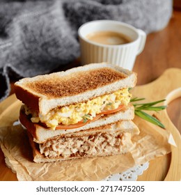 A professional culinary set featuring two types of toasted club sandwiches: a creamy egg salad sandwich with fresh tomato slices and a savory tuna salad sandwich. The golden-brown toasted bread is perfectly layered with rich fillings and garnished with a fresh rosemary sprig. Presented on a rustic wooden pizza-style serving board with white lace paper doilies and brown parchment paper. The collection includes a top-down flat lay and a 45-degree angle close-up shot, accompanied by a cup of hot espresso coffee with rich crema. Set against a wooden tabletop with a cozy grey knitted background. Ideal for cafe menus, breakfast promotion, healthy lunch blogs, and gourmet recipe content. Bright, natural lighting highlights the fresh textures and appetizing presentation.