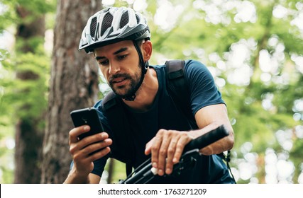 Professional cyclist resting in the forest, holding mobile phone, using online application for searching GPS coordinates while riding the bike. Travel, sport and modern technology concept. - Powered by Shutterstock