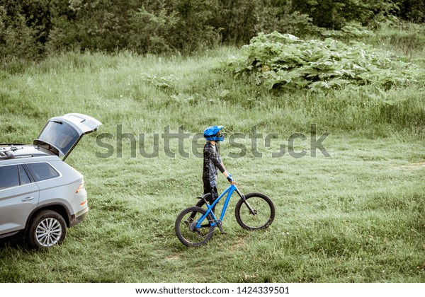 Professional cyclist\
walking with bicycle near his car on the green meadow in the\
mountains. View from\
above