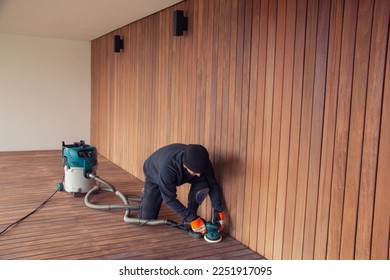 Professional deck worker kneeling while sanding wooden deck with orbital power sander collecting dust to industrial vacuum cleaner - Shutterstock ID 2251917095