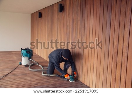 Professional deck worker kneeling while sanding wooden deck with orbital power sander collecting dust to industrial vacuum cleaner