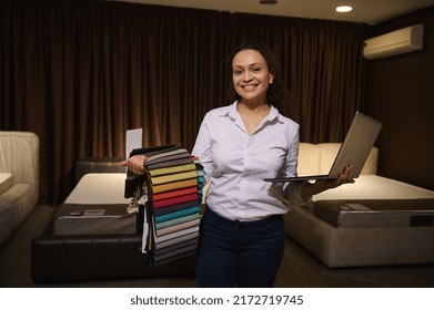 Professional Decorator, Interior Designer Smiles Toothy Smile Looking At Camera, Standing Against Upholstered Beds In A Home Design Showroom, Holding Laptop And Upholstery Fabric Samples In Her Hands
