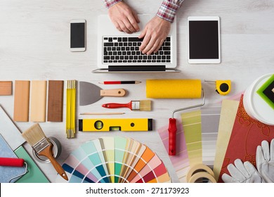 Professional Decorator's Hands Working At His Desk And Typing On A Laptop, Color Swatches, Paint Rollers And Tools On Work Table, Top View